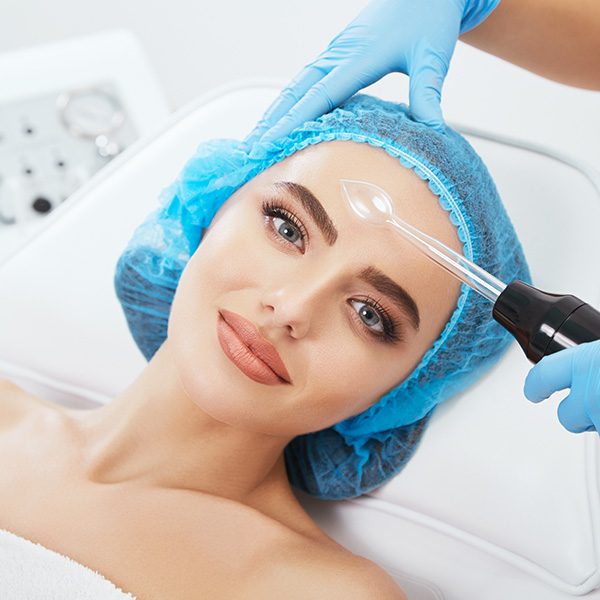 head-shoulders-woman-lying-couch-blue-cap-cosmetological-clinic-looking-camera-doctor-holding-darsonvales-near-her-face